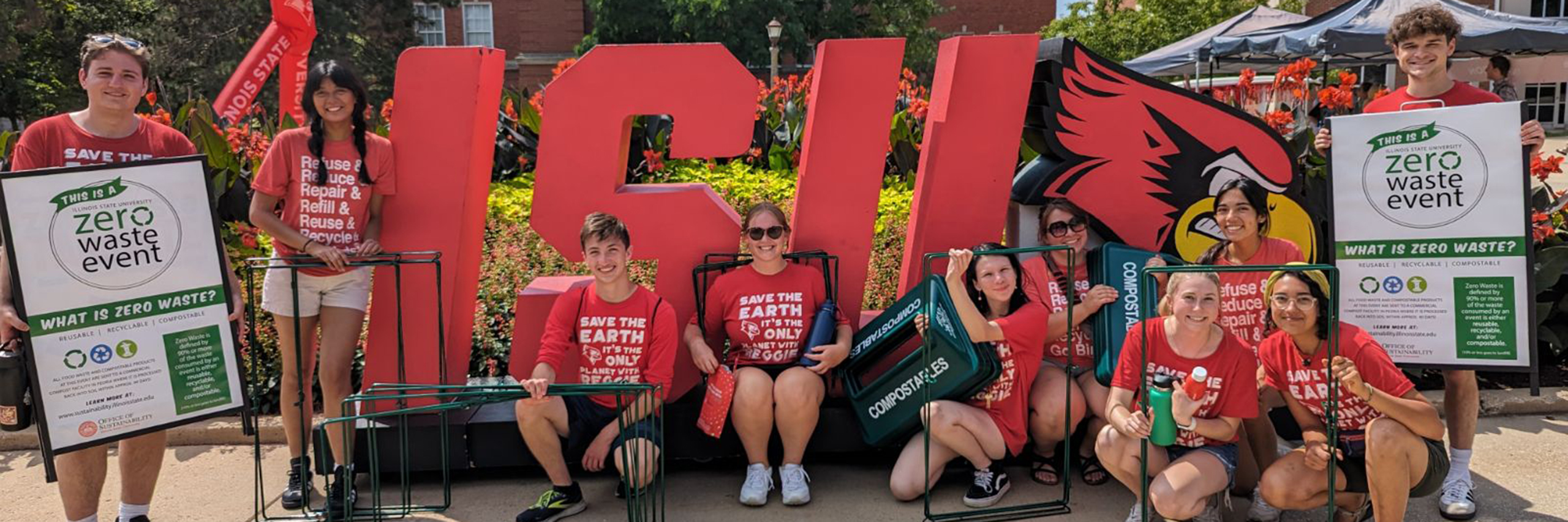 a group of people standing and sitting in front of large red letters spelling ISU. Behind them are green bushes and red flowers. The people are wearing red t-shirts with various environmental slogans and are holding signs and bins for compostables. Two larger signs flank the group, each promoting a Zero Waste Event with text about the event's aims and features.