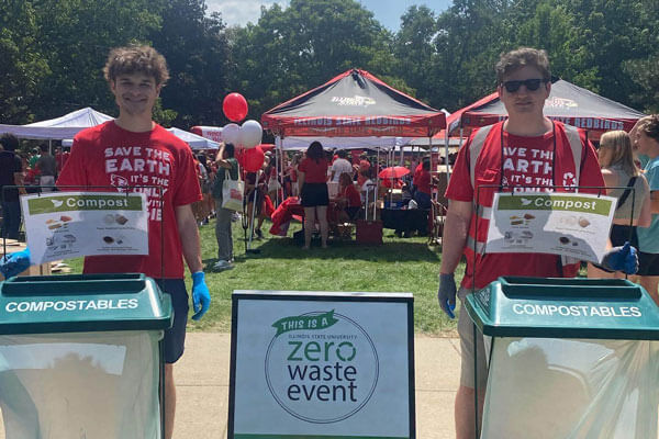Two individuals at a compost station during a zero waste event, standing beside green bins labeled 'COMPOSTABLES.'