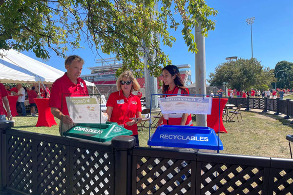Three people stand near compost and recycling bins at an outdoor event, with a stadium and tent in the background.