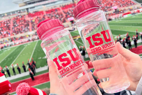 Two clear water bottles with 'ISU ZERO WASTE' in red text being held at a stadium with a football field in the background.