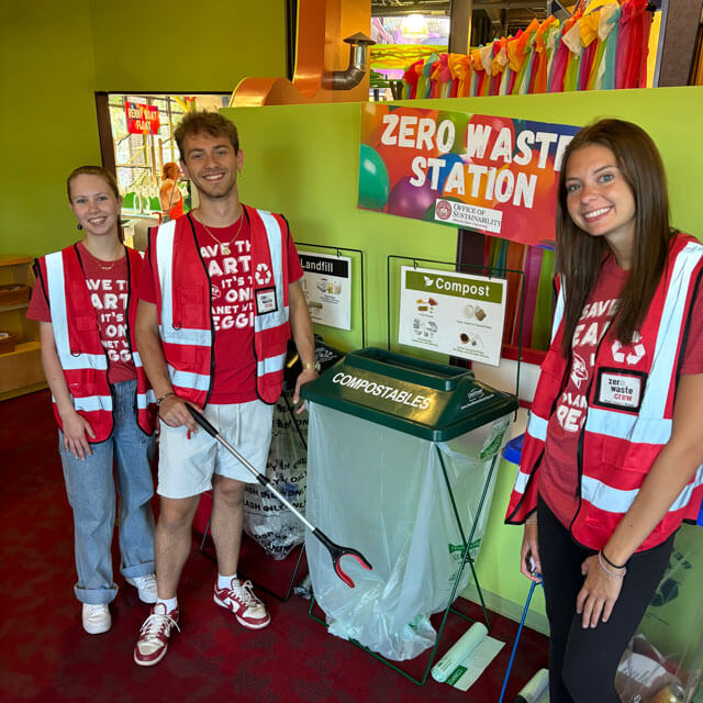 Three people wearing red vests stand beside a Zero Waste Station.