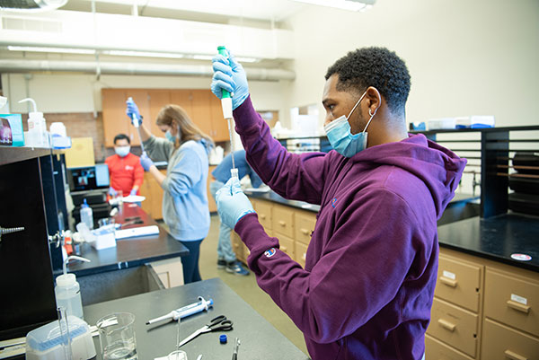 A student, wearing gloves and a mask, drops liquid into a small vial.